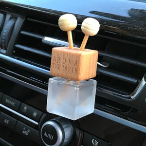 May include: A wooden car air freshener with two wooden sticks and a glass bottle. The bottle is frosted and has the text "Aroma Therapie" on the front. The air freshener is attached to a car's air vent.