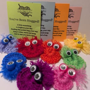 Könnte beinhalten: Ein Set aus acht gehäkelten kuscheligen Spielzeugen mit Glitz Augen. Die Spielzeuge sind in verschiedenen Farben, darunter Lila, Blau, Rot, Orange, Grün und Gelb. Jedes Spielzeug hat ein Etikett mit der Aufschrift "You've Been Hugged!" und enthält ein kurzes Gedicht über das Senden einer Umarmung.