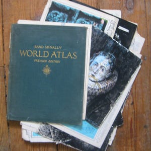 May include: A vintage green hardcover book titled "RAND MCNALLY WORLD ATLAS PREMIER EDITION" with a compass illustration on the cover. The book is stacked on top of other books and maps.