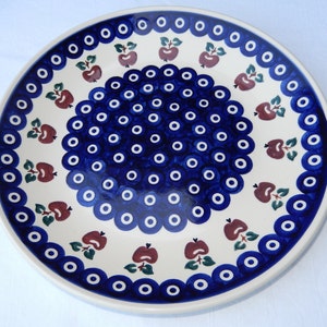 May include: A white ceramic plate with a blue and white pattern of circles and a border of red apples with green leaves.