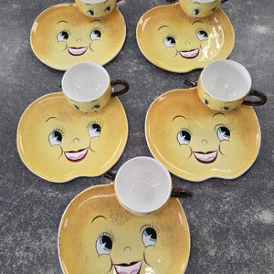 May include: A set of five whimsical teacups and saucers. Each saucer is shaped like a yellow apple with a smiling face design. The teacups are yellow with brown handles and white interiors. The set is arranged on a gray surface.