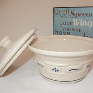 Peut inclure: Plat à four en céramique blanc cassé avec couvercle, orné de motifs floraux et rubans bleus. Le couvercle est légèrement ouvert. Un panneau en bois en arrière-plan indique "Until you spread your wings you will have no idea."