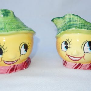 May include: A pair of vintage ceramic kitchenware items. One is a covered container, the other a creamer. Both are yellow with smiling faces, blue eyes, and green hats. They have pink and red scarves.