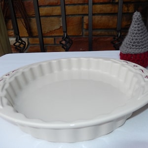 Peut inclure: Un plat à tarte en céramique blanc cassé avec un bord cannelé et un rebord décoratif orné de petits motifs floraux bordeaux. Le plat présente une surface intérieure lisse et brillante ainsi qu'une paroi extérieure festonnée.
