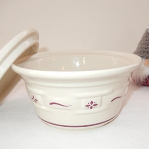 Può includere: Ciotola in ceramica bianco sporco con coperchio, con un motivo floreale decorativo in una tonalità rosso scuro. La ciotola è rotonda con un bordo leggermente svasato. Un piccolo gnomo con cappello grigio e barba bianca sullo sfondo.