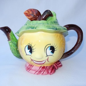 May include: A whimsical, vintage teapot with a smiling face design. The teapot is primarily yellow with a green leafy hat, brown handle and spout, and a pink and red striped scarf. The face has blue eyes and long eyelashes.