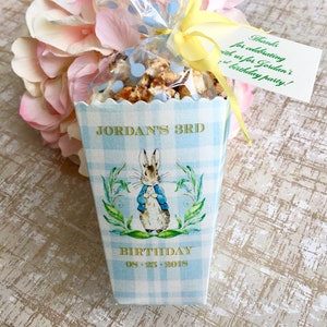 May include: A blue and white checkered favor box with a Peter Rabbit illustration. The box has gold lettering that says "Jordan's 3rd Birthday 08.25.2018".