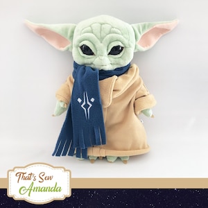 May include: A plush doll of a character with large ears, green skin, and large black eyes. The doll wears a tan coat and a blue scarf with a white emblem. The doll is a Star Wars character.
