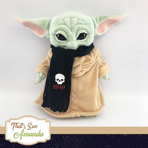 May include: A plush doll of a character with large ears, wearing a tan robe and a black scarf with a white skull design. The doll has large black eyes and green skin. The image includes the text "That's Sew Amanda".