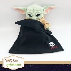 May include: A plush green and brown toy character with large ears, wearing a black fleece blanket with a white skull and red crossbones embroidered on it. The blanket has a fold-over design and is tucked around the toy character.