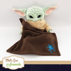 Puede incluir: Una manta de franela marr&oacute;n con una rana azul bordada en ella. Un juguete de peluche verde est&aacute; parcialmente cubierto por la manta. El texto "That's Sew Amanda" est&aacute; en una etiqueta en la esquina inferior izquierda de la imagen.