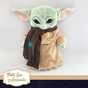 May include: A plush toy of a green character wearing a brown scarf and a tan coat. The character has large eyes and a small mouth. The scarf has a blue frog embroidered on it. The text "That's Sew Amanda" is visible in the bottom left corner of the image.
