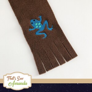 May include: Brown fleece scarf with a blue and green embroidered frog design. The scarf has fringe on the bottom.