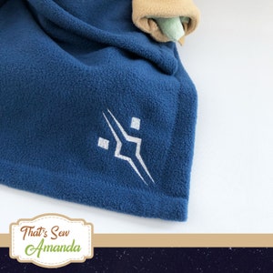 May include: A blue fleece blanket with a white embroidered logo. The logo is a stylised geometric design with a square in the centre and two triangles extending out from the square. The blanket is folded and the logo is visible in the centre of the image. The text "That's Sew Amanda" is visible in the bottom right corner of the image.