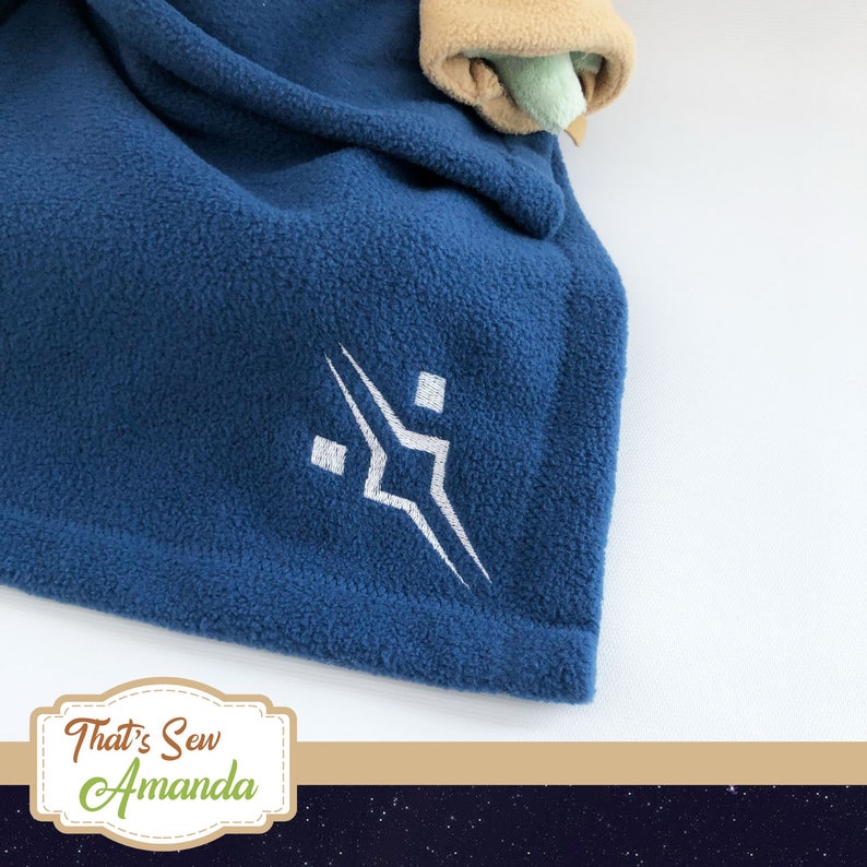 May include: A blue fleece blanket with a white embroidered logo. The logo is a stylised geometric design with a square in the centre and two triangles extending out from the square. The blanket is folded and the logo is visible in the centre of the image. The text "That's Sew Amanda" is visible in the bottom right corner of the image.