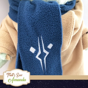 May include: A close-up of a blue fleece scarf with a white embroidered design. The scarf is draped over a tan-colored garment. The scarf has a soft, textured appearance. The image includes the text "That's Sew Amanda".