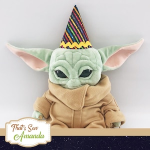 May include: A plush toy of a green alien character wearing a colorful party hat. The alien has large ears and big eyes. The toy is wearing a brown, furry coat.