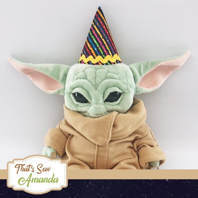 May include: A plush toy of a green alien character wearing a colorful party hat. The alien has large ears and big eyes. The toy is wearing a brown, furry coat.