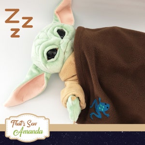 Puede incluir: Un juguete de peluche verde con ojos grandes, que lleva una manta marr&oacute;n con una rana azul bordada en ella. El juguete est&aacute; acostado de lado con la cabeza apoyada en la manta. El texto "That's Sew Amanda" es visible en la esquina inferior izquierda de la imagen.