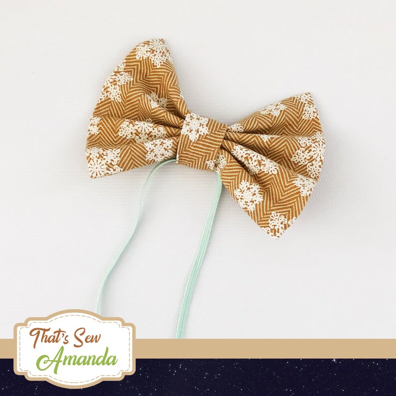 May include: A brown bow tie with a white snowflake pattern and a light green elastic band. "That's Sew Amanda" is written on a label.