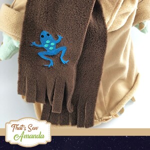 May include: A brown fleece scarf with a blue frog embroidered on it. The scarf has fringe on the bottom and is attached to a tan hooded garment.