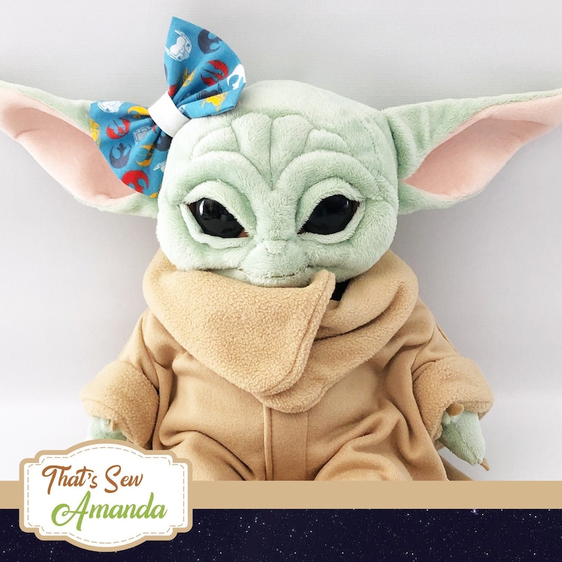 May include: A plush toy of a green character with large eyes and a brown hooded robe. The character is wearing a blue bow with a white and red pattern. The toy is sitting on a dark background with stars.