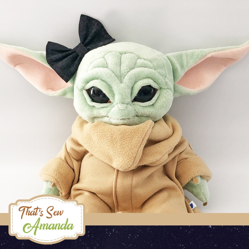 May include: A plush toy of a green character with large ears, wearing a brown coat and a black bow. The toy has large eyes and a serious expression. The text "That's Sew Amanda" is visible in the lower left corner of the image.