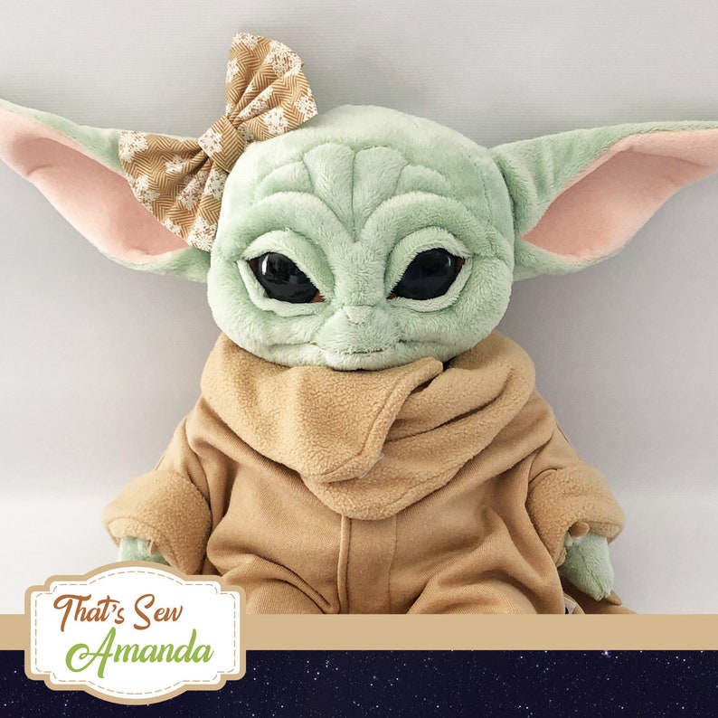 May include: A plush toy of a green character with large eyes, wearing a brown hooded robe and a brown and gold bow. The toy is sitting on a dark blue background with white stars. The text "That's Sew Amanda" is visible in the lower left corner of the image.