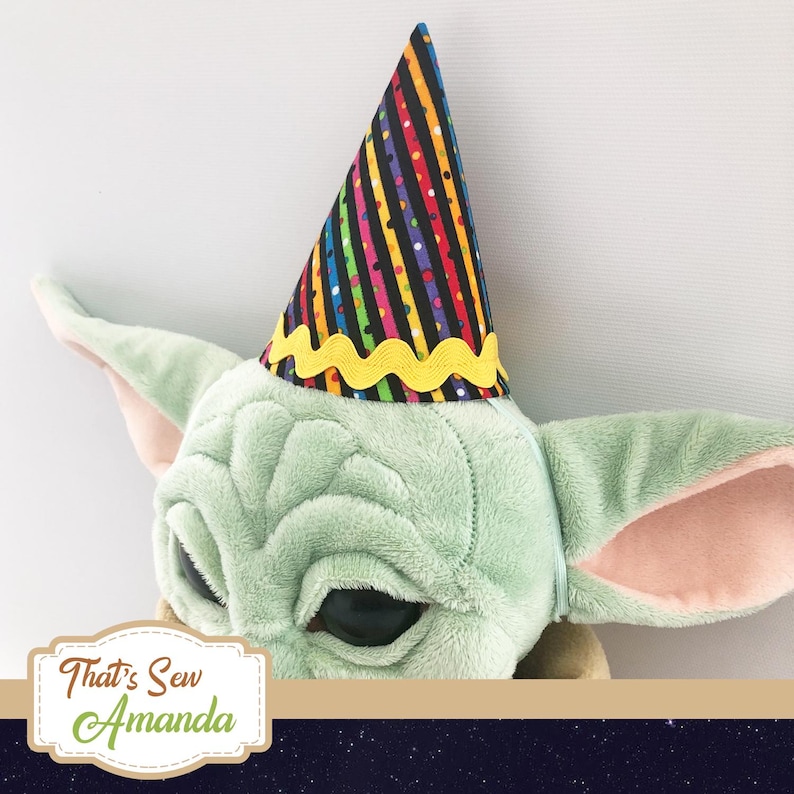 May include: A green plush toy with large ears wearing a black and rainbow striped party hat with a yellow zig-zag trim. The hat has black polka dots and colorful stripes. The toy has large black eyes and a small mouth. The text "That's Sew Amanda" is visible in the bottom left corner of the image.