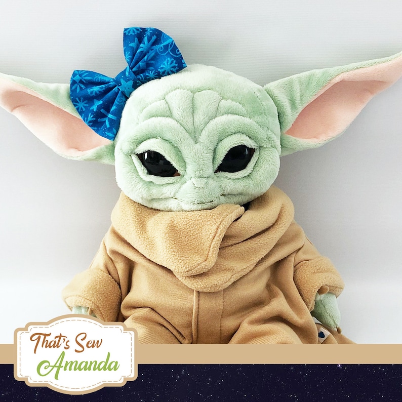 May include: A plush toy of a green character with large eyes and a brown coat. The character is wearing a blue bow with a snowflake pattern. The toy is sitting on a dark blue background with white stars.