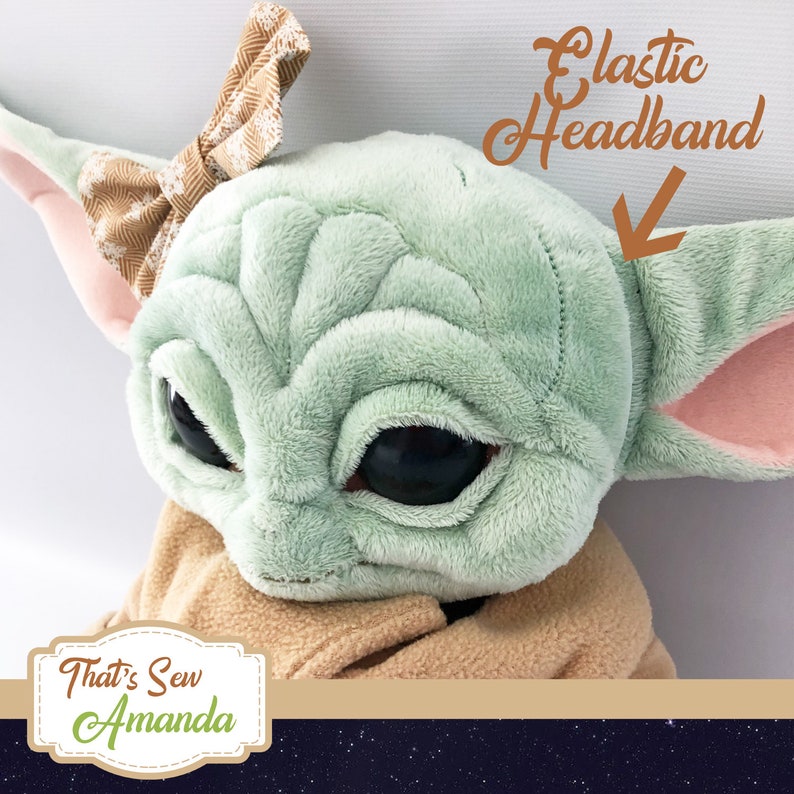 May include: A green plush toy with large eyes and a brown bow on its head. The toy is wearing a brown plush robe and has an elastic headband.