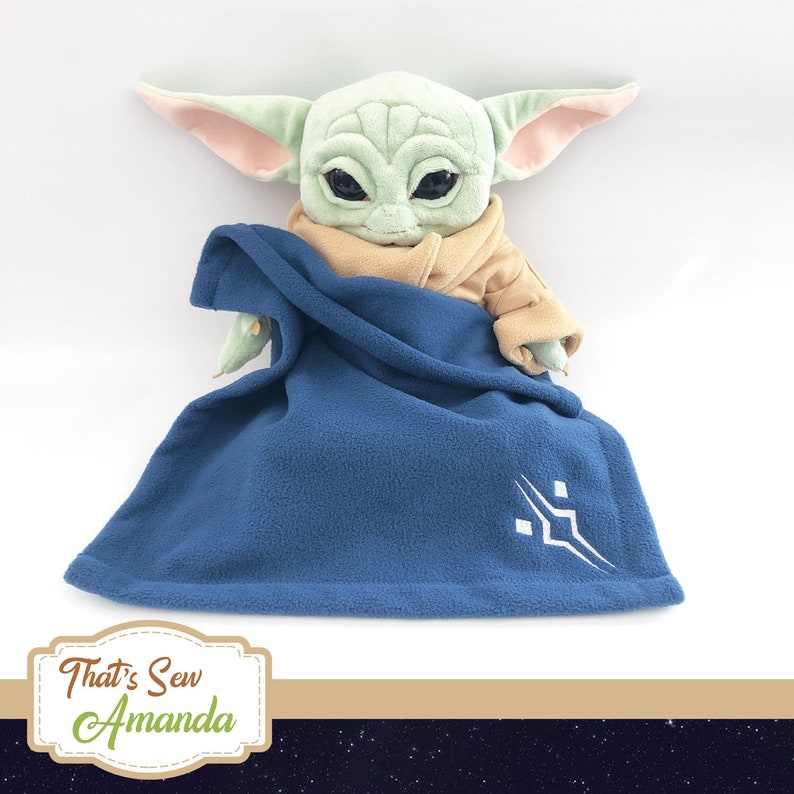 May include: A plush toy of a green baby alien character, wearing a tan robe and a blue blanket with a white symbol. The toy is sitting on a white background.