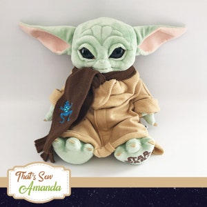 May include: A green plush toy with large ears, brown eyes, and a brown scarf. The toy is wearing a tan coat with a brown frog embroidered on the scarf. The toy is sitting on a white surface. The text "Star Wars" is visible on the toy's foot.