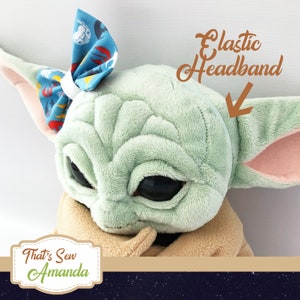 May include: A plush toy of a green character with large eyes and a brown robe. The character is wearing a blue and red bow headband with an elastic band.