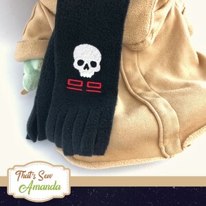 May include: Black scarf with a white skull and red design. The scarf is fringed at the bottom and is draped over a tan hooded garment. The scarf has a skull and the number 22 embroidered on it.