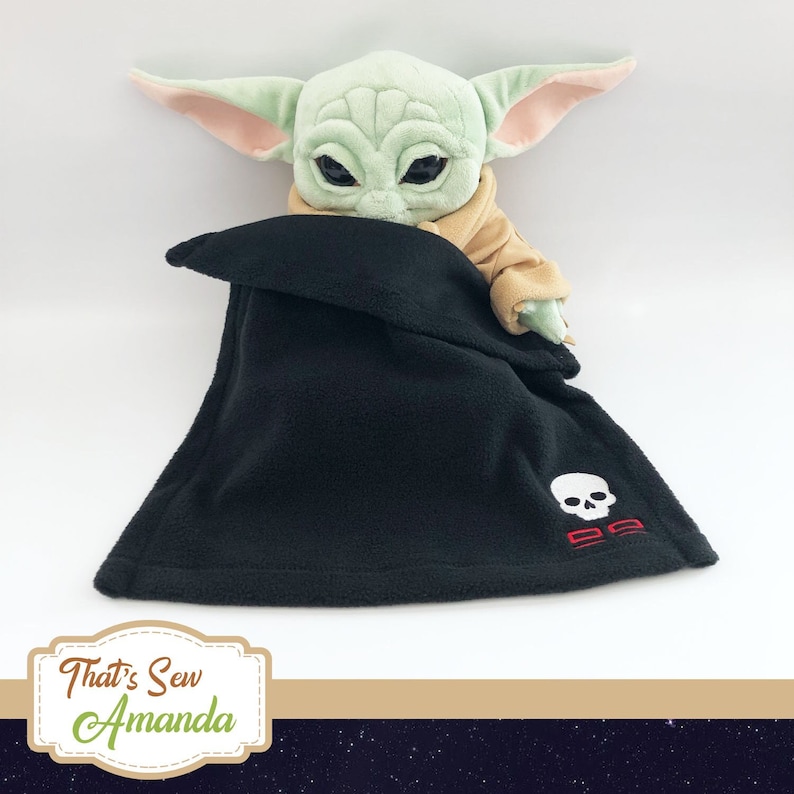 May include: A plush green and brown toy character with large ears, wearing a black fleece blanket with a white skull and red crossbones embroidered on it. The blanket has a fold-over design and is tucked around the toy character.