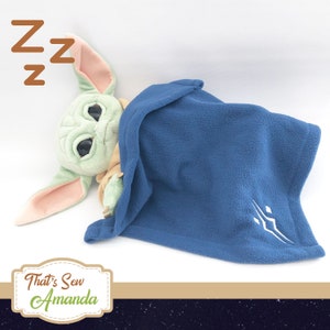 May include: A green plush toy with large eyes, resembling a baby, is tucked into a blue fleece blanket with a white embroidered symbol. The toy is lying on a white surface with the text "That's Sew Amanda" in a brown frame below.