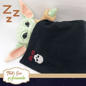 May include: A green plush toy with large eyes is sleeping under a black fleece blanket with a skull and crossbones design embroidered on it. The blanket has the text "55" embroidered in red above the skull. The toy is holding a green pear-shaped object in its hand.