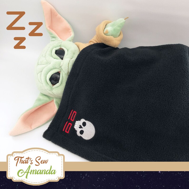May include: A green plush toy with large eyes is sleeping under a black fleece blanket with a skull and crossbones design embroidered on it. The blanket has the text "55" embroidered in red above the skull. The toy is holding a green pear-shaped object in its hand.