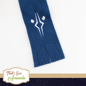 May include: A navy blue fleece scarf with a white embroidered geometric design. The scarf has a fringed bottom edge. The scarf is a cozy accessory.