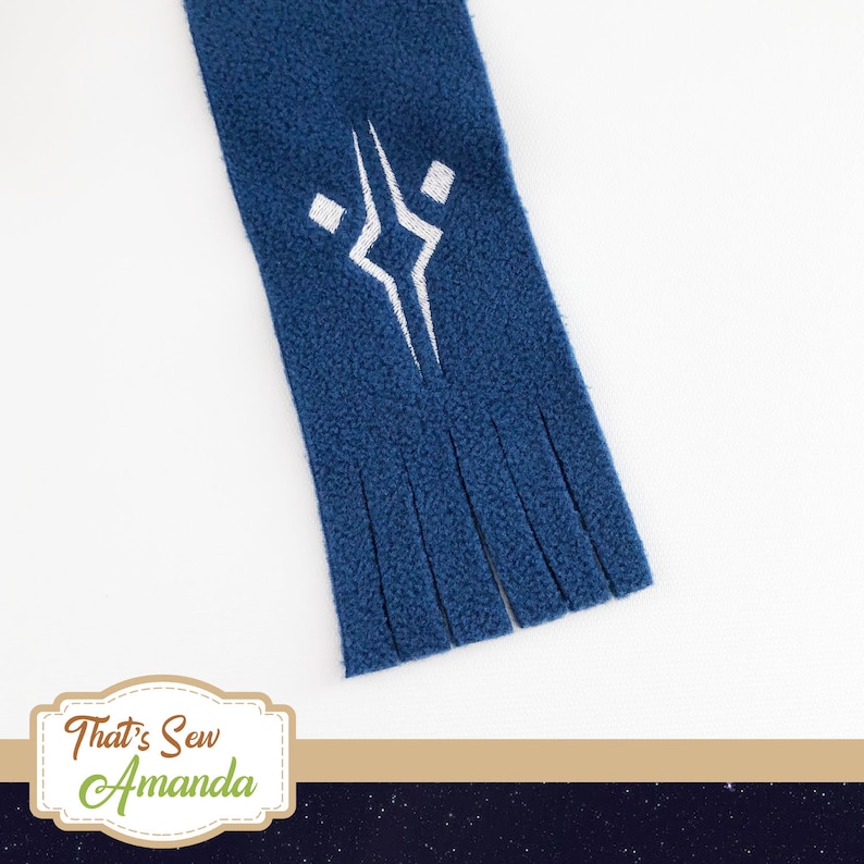 May include: A navy blue fleece scarf with a white embroidered geometric design. The scarf has a fringed bottom edge. The scarf is a cozy accessory.