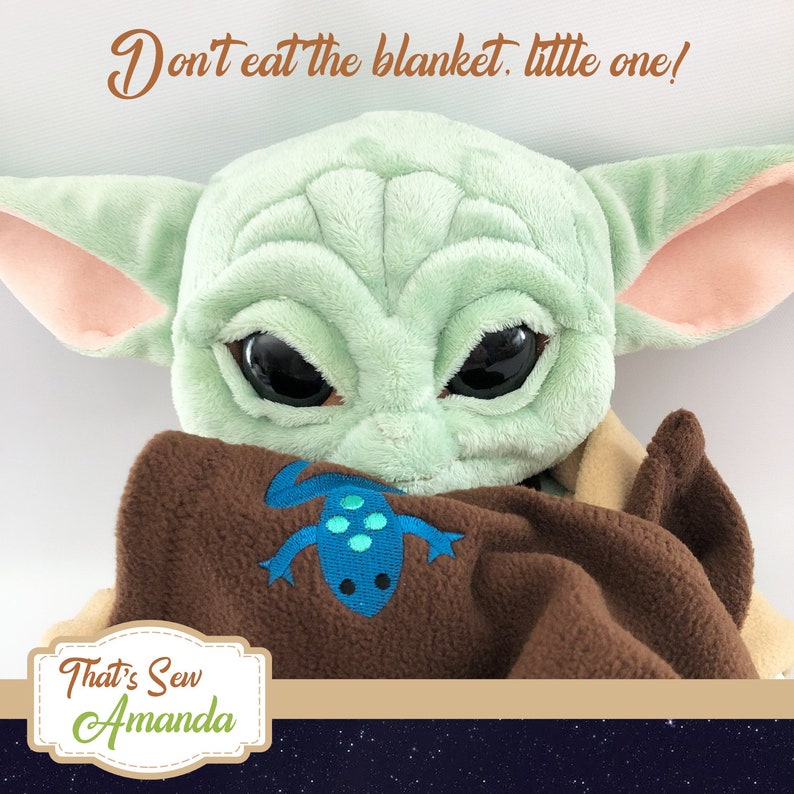 Puede incluir: Un juguete de peluche verde con ojos grandes, que lleva una manta marr&oacute;n con un dise&ntilde;o de lagarto azul y verde. El juguete est&aacute; sujetando la manta y el texto "No te comas la manta, peque&ntilde;o!" est&aacute; escrito encima del juguete. El texto "That's Sew Amanda" est&aacute; escrito en una etiqueta en la esquina inferior izquierda de la imagen.