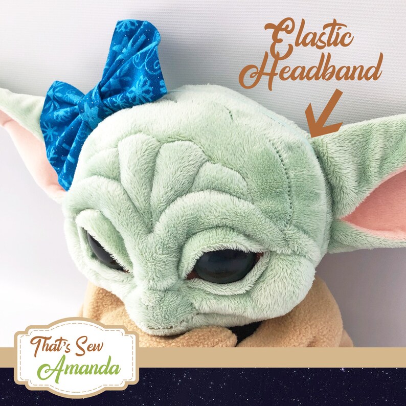 May include: A plush toy of a green character with large eyes, wearing a blue bow headband. The headband is elastic and has a snowflake pattern. The toy is sitting on a brown surface. The text "That's Sew Amanda" is visible in the lower left corner of the image.