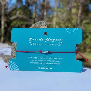 Puede incluir: Una pulsera de hilo rojo con tres cuentas plateadas se muestra sobre una tarjeta turquesa. La tarjeta tiene el texto "Rio de Magma" y "JOYERÍA ARTESANAL". La tarjeta también incluye texto en español sobre las propiedades protectoras de la pulsera.
