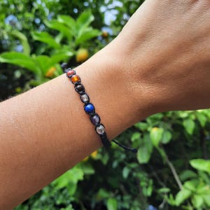 May include: A black macrame bracelet featuring seven colorful gemstone beads. The beads are arranged in a row, with colors including red, orange, brown, clear, blue, purple, and clear. The bracelet is worn on a wrist.