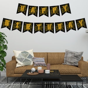 Puede incluir: Una pancarta negra con letras doradas que dice "HAPPY BIRTHDAY" cuelga de una pared blanca. La habitación cuenta con un sofá marrón, una mesa de centro negra y una gran planta verde. La pancarta es un elemento decorativo para una celebración.