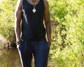 May include: A person wearing a black tank top with a silver pendant necklace and dark blue pants. They are standing outdoors near a body of water and greenery.