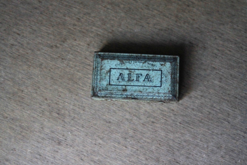 Peut inclure: Une bo&icirc;te vintage en m&eacute;tal bleu avec une bordure blanche et le mot "ALFA" imprim&eacute; en lettres noires.