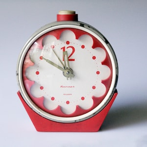 Puede incluir: Un reloj despertador analógico vintage rojo y blanco con una esfera festoneada. El reloj tiene una base roja y un marco plateado. La esfera muestra el número 12 en rojo y tiene marcadores de hora de puntos rojos.