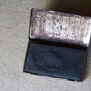 Peut inclure: Une petite bo&icirc;te rectangulaire en m&eacute;tal avec un couvercle &agrave; charni&egrave;re. La bo&icirc;te est ouverte, r&eacute;v&eacute;lant un bloc rectangulaire noir &agrave; l'int&eacute;rieur. La bo&icirc;te a une finition argent&eacute;e us&eacute;e.